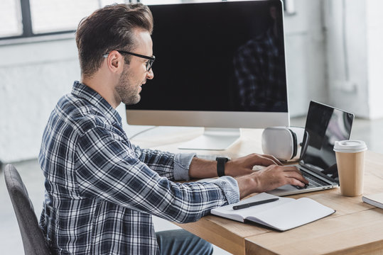 side view of smiling young man in eyeglasses using laptop at workplace