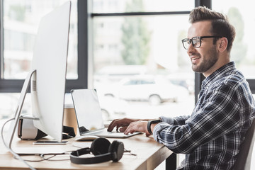 smiling young man in eyeglasses using laptop and desktop computer at workplace