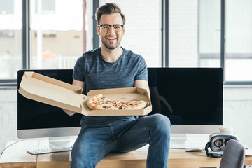 handsome young programmer in eyeglasses holding pizza and smiling at camera