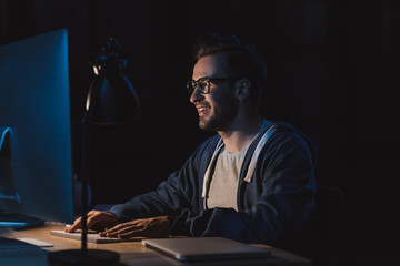 smiling young programmer in eyeglasses working with desktop computer at night