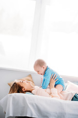 side view of mother playing with laughing little child while lying on bed at home