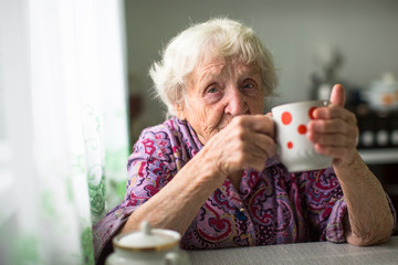 An elderly woman drinks tea sitting in the kitchen.