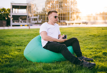 Handsome young hipster man sitting on soft chair with smartphone in hands outdoors in park