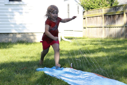 Little Preschool Girl Runs Through A Backyard Slip And Slide Water Slide. A Fun Backyard Activity For Young Children, Shows Themes Of Summer And Backyard Fun