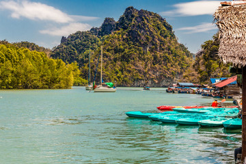 Beautiful scenery of two sailing boats next to a mangrove coastline and many moored kayaks and motorboats at a floating platform with a limestone formation in the background; in Kilim Geoforest Park.
