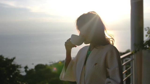 Young beautiful woman wears bathrobe drinks coffee in the morninig during amazing sunrise stands on the terrace of her villa enjoying the moment. slow motion. 3840x2160, 4k
