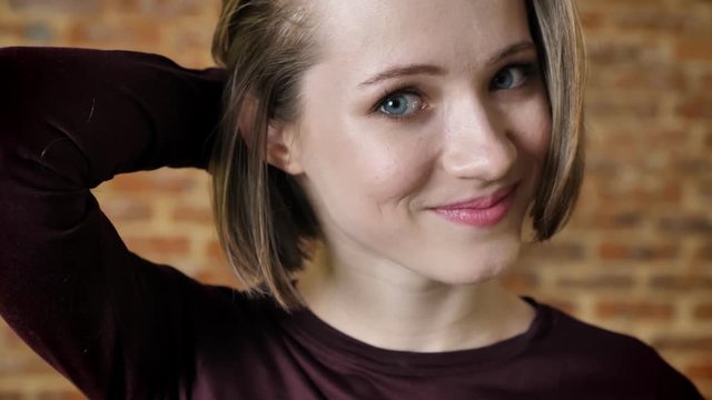 Young attractive shy girl is smiling, look away, tucking hair, watching at camera, brick background