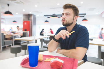 Portrait of a man with French fries in his arms sitting in a bright fast food restaurant and eating fast food. Student sits at a table with a tray on which a cool drink, a burger and French fries.