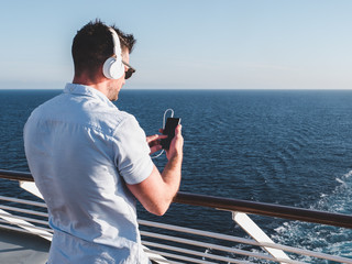 Stylish, attractive guy in sunglasses with phone and white headphones on the deck of the cruise ship on the background of the morning sunrise and blue sky. Concept of sea travel and recreation