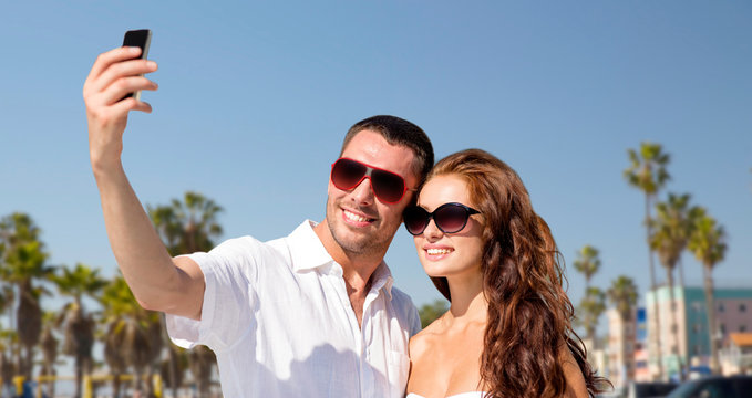 travel, tourism and technology concept - smiling couple in sunglasses making selfie by smartphone over venice beach background in california