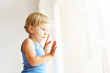 Fototapeta premium Child missing his mother concept. Little cute blond three year old boy sitting on windowsill touching window glass, looking sad. Kid in blue tank top playing alone. Background, close up, copy space.