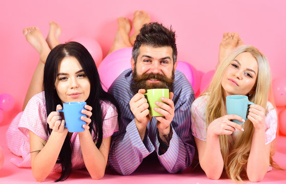 Man and women, friends on sleepy faces lay, pink background. Lovers drinking coffee in bed. Threesome relax in morning with coffee. Stormy night concept. Man and women in domestic clothes, pajamas.