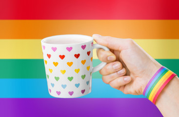 homosexual and lgbt concept - close up of female hand with cacao drink in cup with heart pattern and gay pride awareness wristband over rainbow background