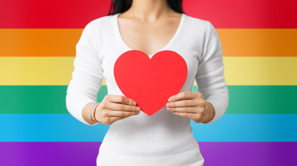 homosexual and lgbt concept - woman holding red heart shape and wearing gay pride awareness ribbon wristband over rainbow background