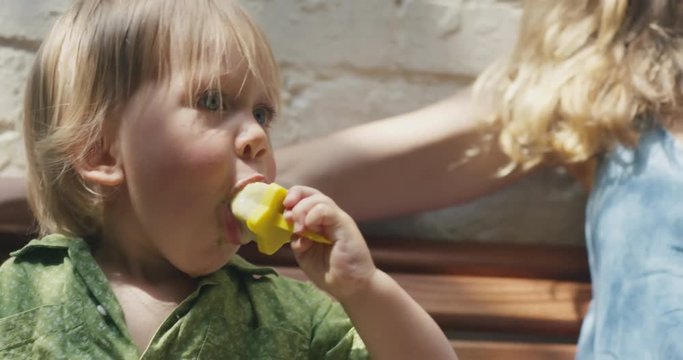 Little toddler boy on bench with mother eating ice lollies