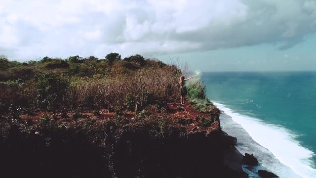 Aerial drone shot of pretty girl playing  with colored smoke on the cliff with amazing ocean and cloudy sky view - video in slow motion