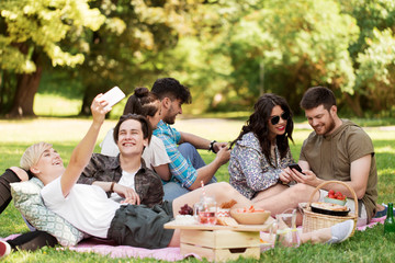 friendship, leisure, technology and people concept - group of friends with smartphones and food chilling on picnic blanket at summer park