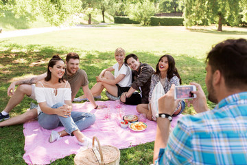 friendship, leisure and technology concept - man taking picture of his friends by smartphone on picnic at summer park