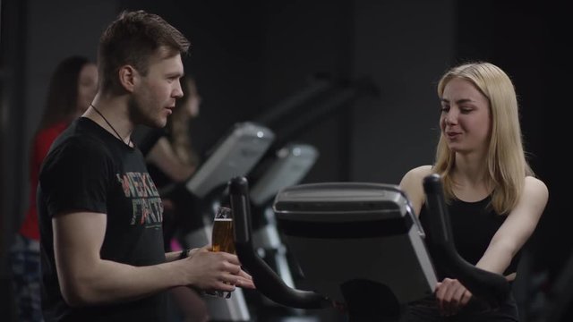 lovely girl in blak sport wear vigorously works on exercise bike and guy comes up with a glass of beer and offers her a drink in the new gym against the background of engaged girls