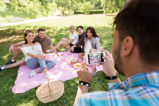 friendship, leisure and technology concept - man taking picture of his friends by smartphone on picnic at summer park