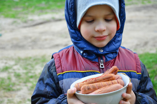 Cute little child, blonde toddler boy with closed eyes biting on sausage from metal fork nearby barbecue area, summer outdoor