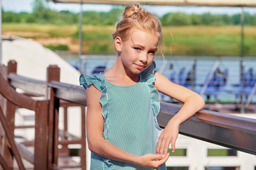 Portrait elegant young girl of 9-11 years outdoors, leaning her elbows on the railing. Cute child blonde smiling, sunny summer day resting on pier. Fashion kid concept. striped dress, marine style.