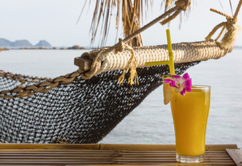 Mango juice decorated with pineapple slice, pink flower and yellow straw next to net hammock by the...