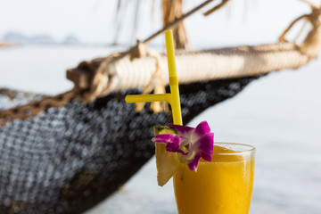 Mango juice close up with pineapple slice, pink flower, yellow straw and beach hammock by the sea...