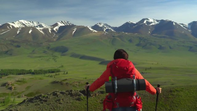 The woman with a red backpack costs on a slope of the mountain and looks at snow mountains.