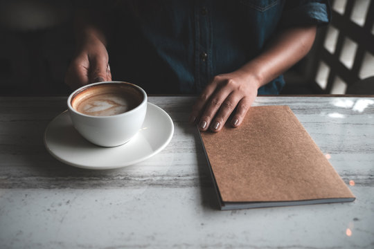 Closeup image of a woman holding and drinking coffee with a notebook on table in cafe