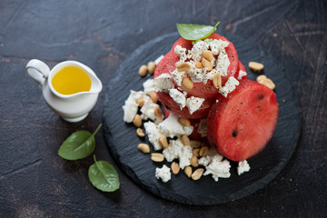 Round slices of watermelon with feta cheese, nuts and olive oil on a stone slate tray, brown stone background, horizontal shot