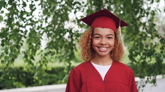 Portrait of pretty African American girl graduating student in red gown and mortarboard standing outdoors, smiling and looking at camera. Youth and education concept.
