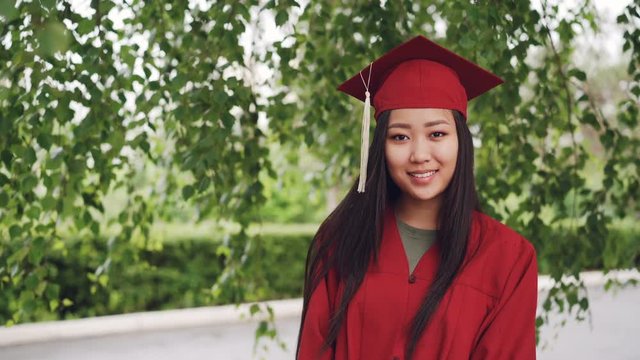 Portrait of attractive Asian girl successful graduating student in gown and mortar-board standing on campus, smiling and looking at camera. Youth and education concept.