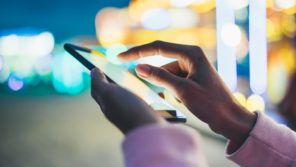 Girl pointing finger on screen smartphone on background bokeh light in night city illumination in evening street defocus, hipster using in hands mobile phone, lifestyle online wifi internet concept