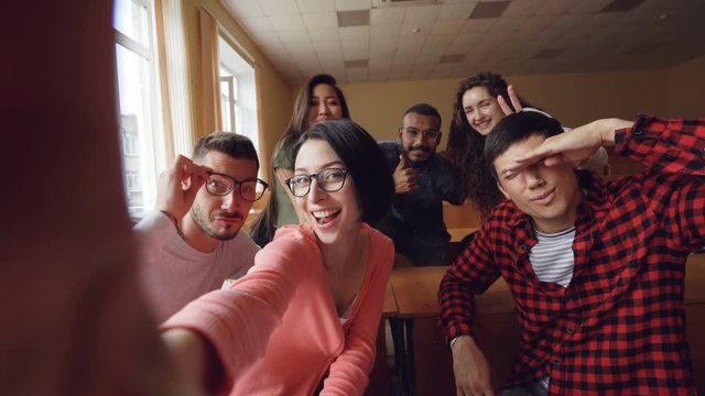Point of view shot of attractive young men and women taking selfie in lecture hall, posing with textbook and showing hand gestures and expressive faces.
