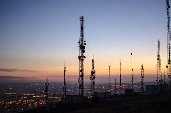 communication tower. cell, radio and television antennas on top of a mountain and below a lit coastal village