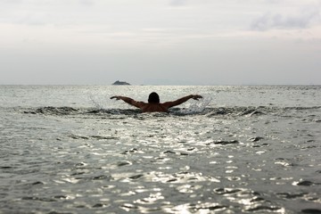 Naklejka premium Silhouette of young man swimming away on butterfly stroke technique towards a small island on the background at sunset in Koh Phangan, Thailand. Hard to reach, destination challenge, effort concepts
