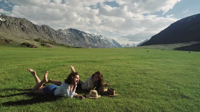 Trendy hipster girls relaxing on the grass. Portrait of a summer lifestyle of two female hipsters lying on the grass of the mountains, enjoying a pleasant day, wearing bright sunglasses. The girl's