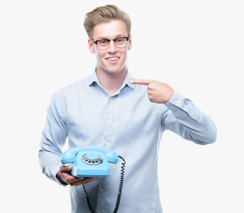 Young handsome blond man holding vintage telephone with surprise face pointing finger to himself