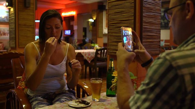 A man takes pictures of a woman eating a roast scorpion and drinking beer in a Thai restaurant