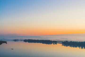 Fototapeta premium Broad mystical river flows along diagonal shore with silhouette of trees and thick fog. Gold glow in predawn sky. Calm morning atmospheric landscape of majestic nature in warm tones.