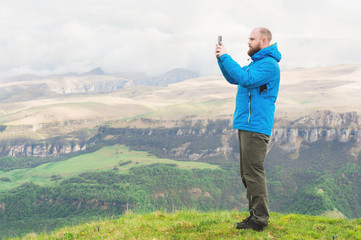 A bearded man in a membrane blue jacket stands in the nature against the backdrop of the mountains and takes pictures on the telephone landscape