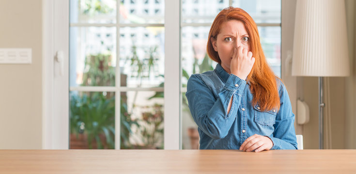 Redhead woman at home smelling something stinky and disgusting, intolerable smell, holding breath with fingers on nose. Bad smells concept.