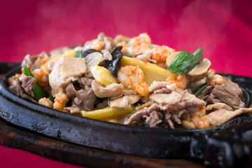 Close up on smokey meat sizzler with prawns, baby corn and mushrooms on iron black plate with wooden base on red tablecloth background. Traditional Chinese hot menu dish