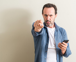 Senior man using smartphone pointing with finger to the camera and to you, hand sign, positive and...