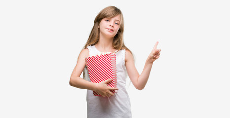 Young blonde toddler holding popcorn pack very happy pointing with hand and finger to the side
