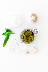 Green pesto sauce in glass jar near basil leaves and garlic on white background top view copy space
