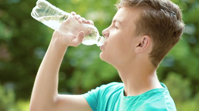 Happy teen boy drinking water from plastic bottle in summer park. Cute teenager drinks cold water - outdoors portrait. 