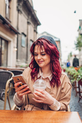 Beautiful young woman having coffee in outdoor cafe while using smartphone. Portrait of stylish girl