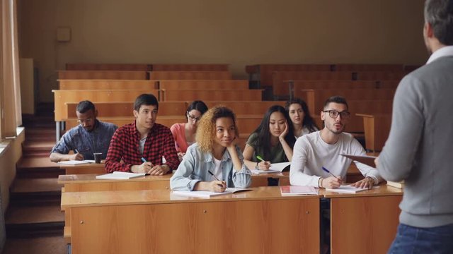 Multi-ethnic group of students is answering teacher's questions, smart young people are talking and smiling while teacher is asking them checking knowledge.